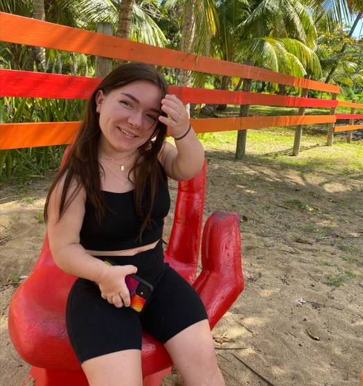 Girl sitting on a red chair shaped like a hand in front of an orange fence and palm trees.